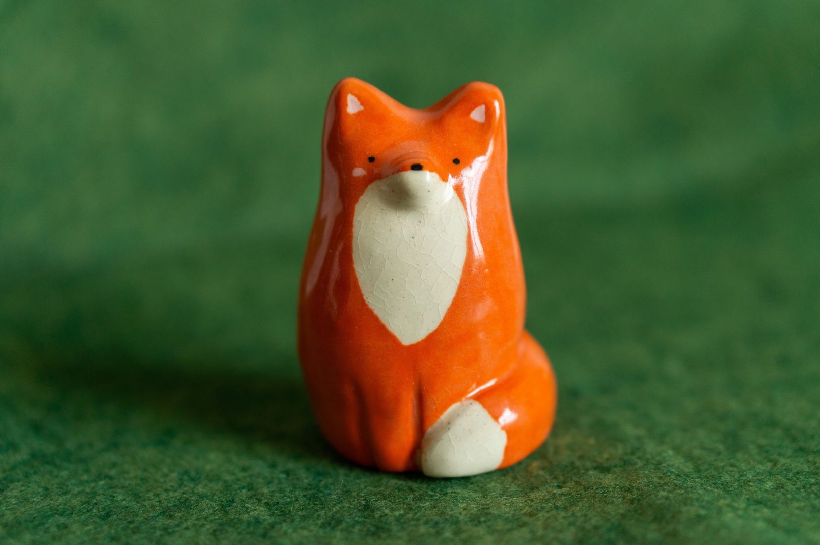 Ceramic fox figurine, about 4 inches tall, in a seated position. Its shape is minimalistic, with smooth rounded edges and a glossy finish. The fox is reddish-orange with a white belly, and has simplistic features including black eyes, a nose, and pink cheeks.