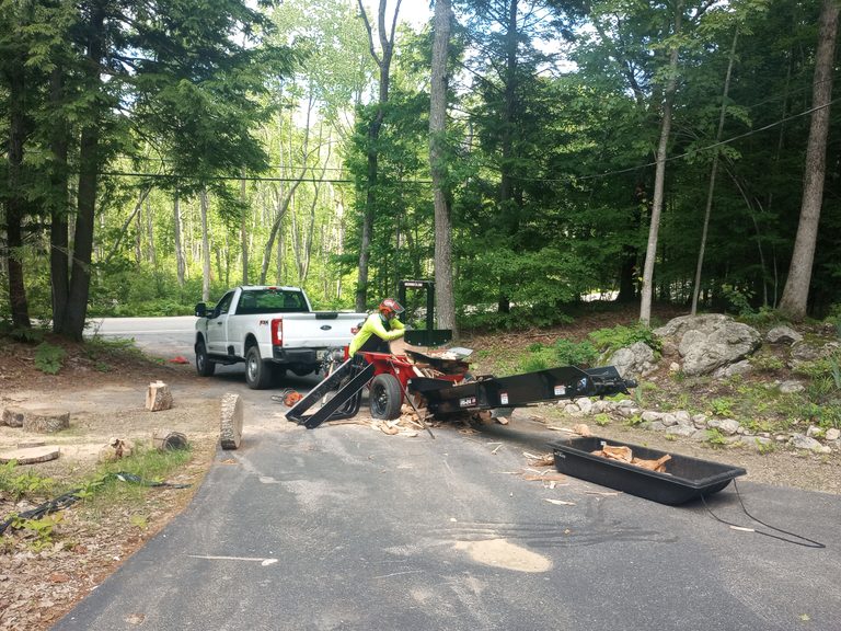 Truck, trailer, and log splitter set up beside a wooded driveway