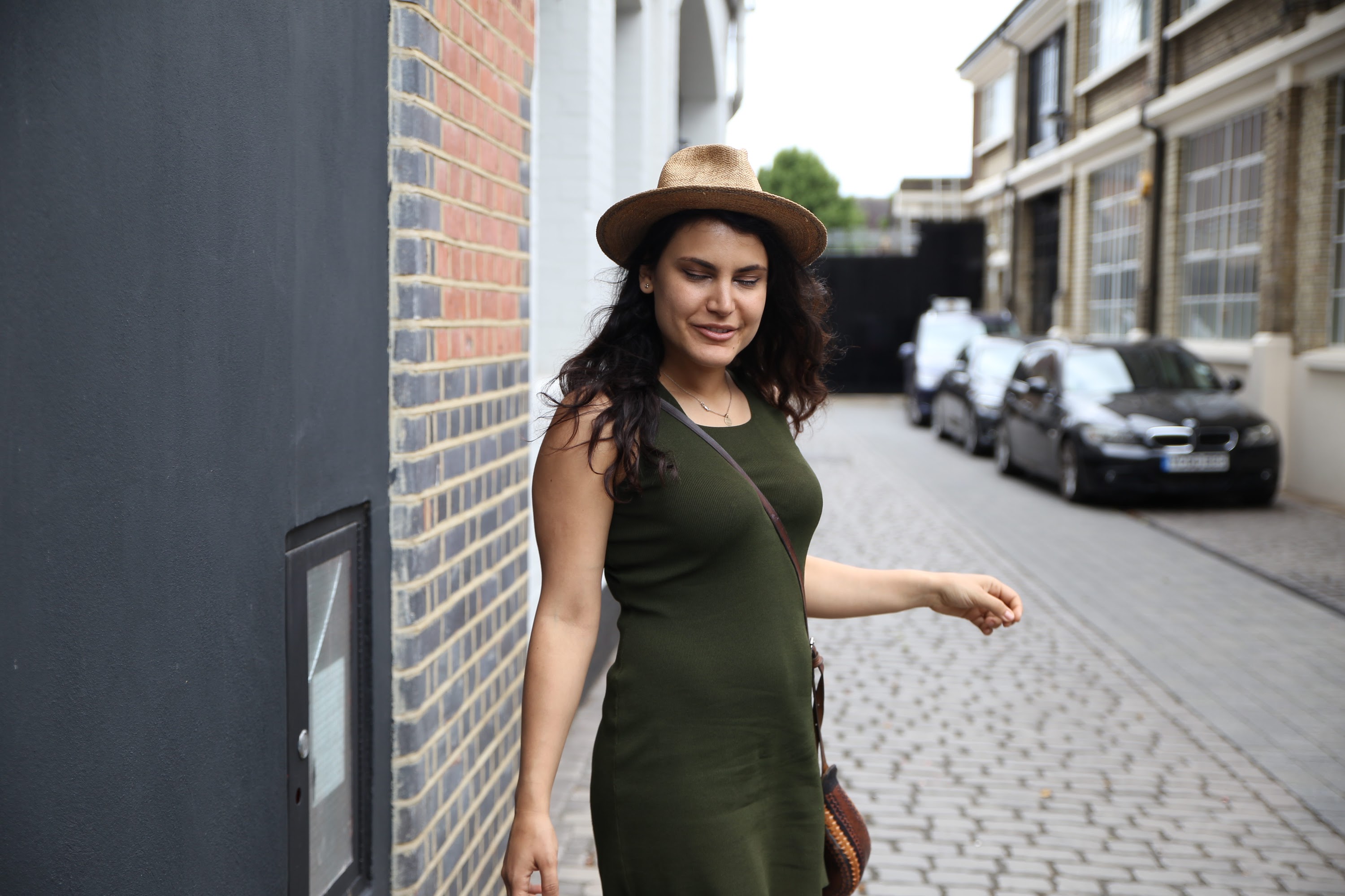 Woman in Green Dress and Straw Hat Walking Down a Cobblestone Street