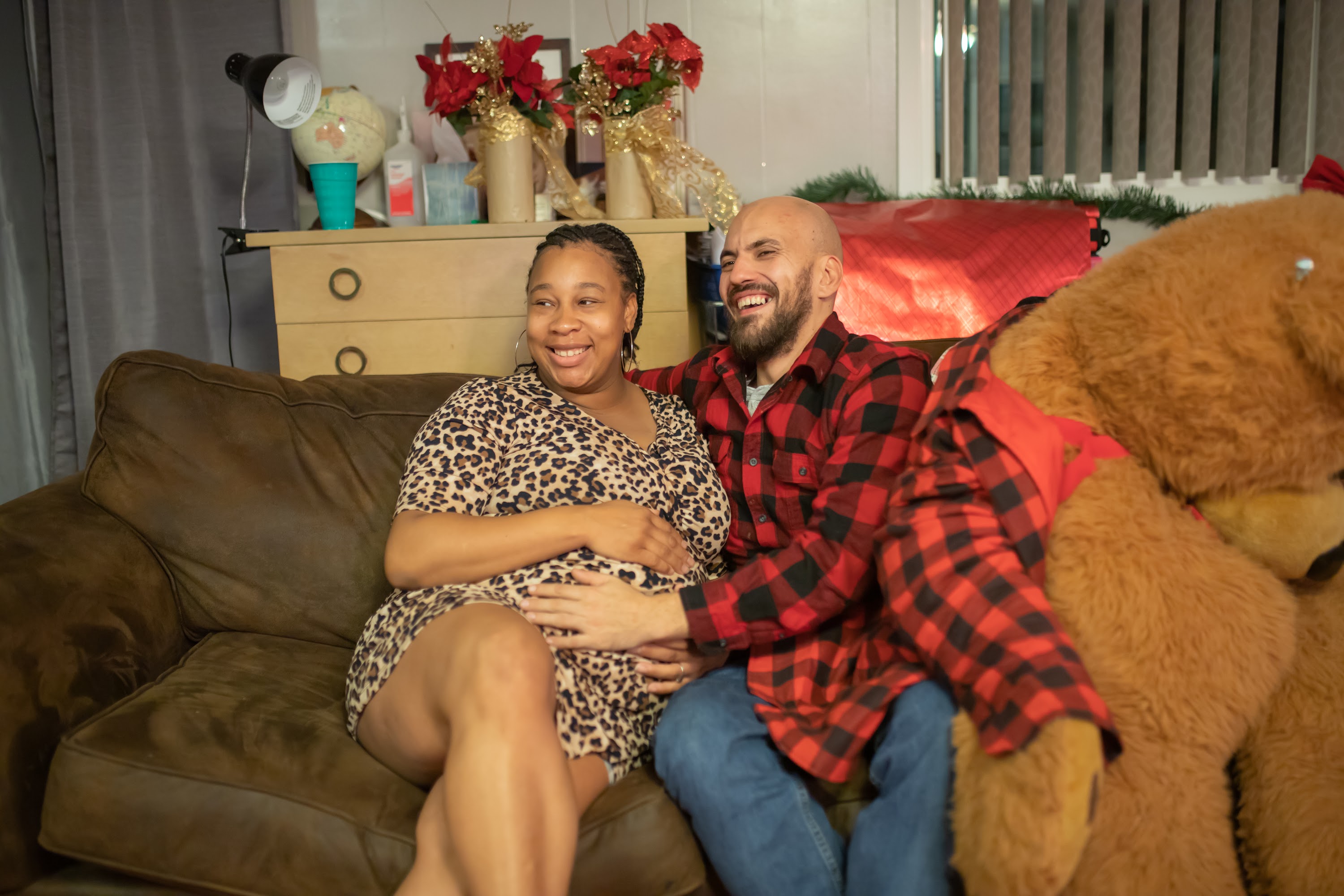 Pregnant Woman and Partner Laughing on Couch with Giant Teddy Bear