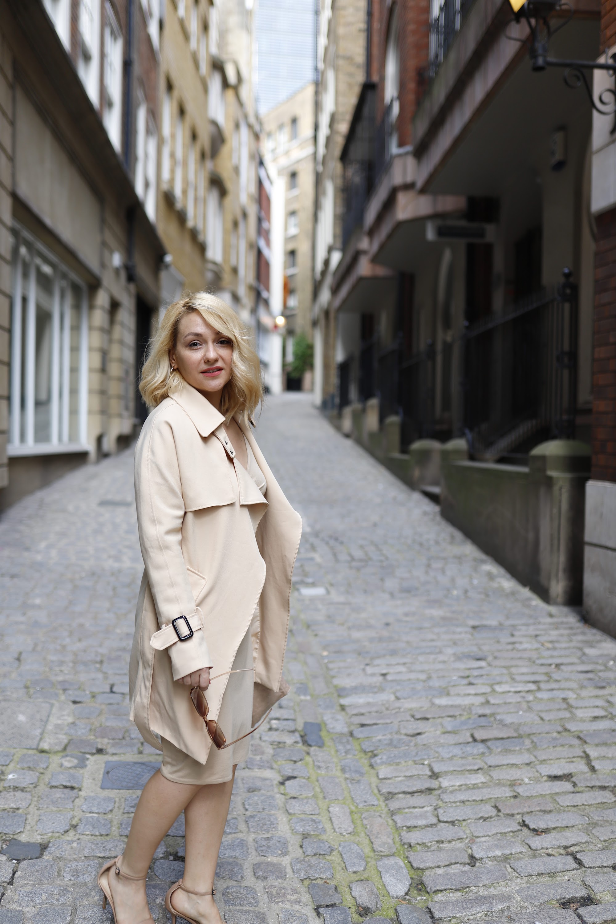 Blonde Woman in Beige Trench Coat on Cobblestone Alley