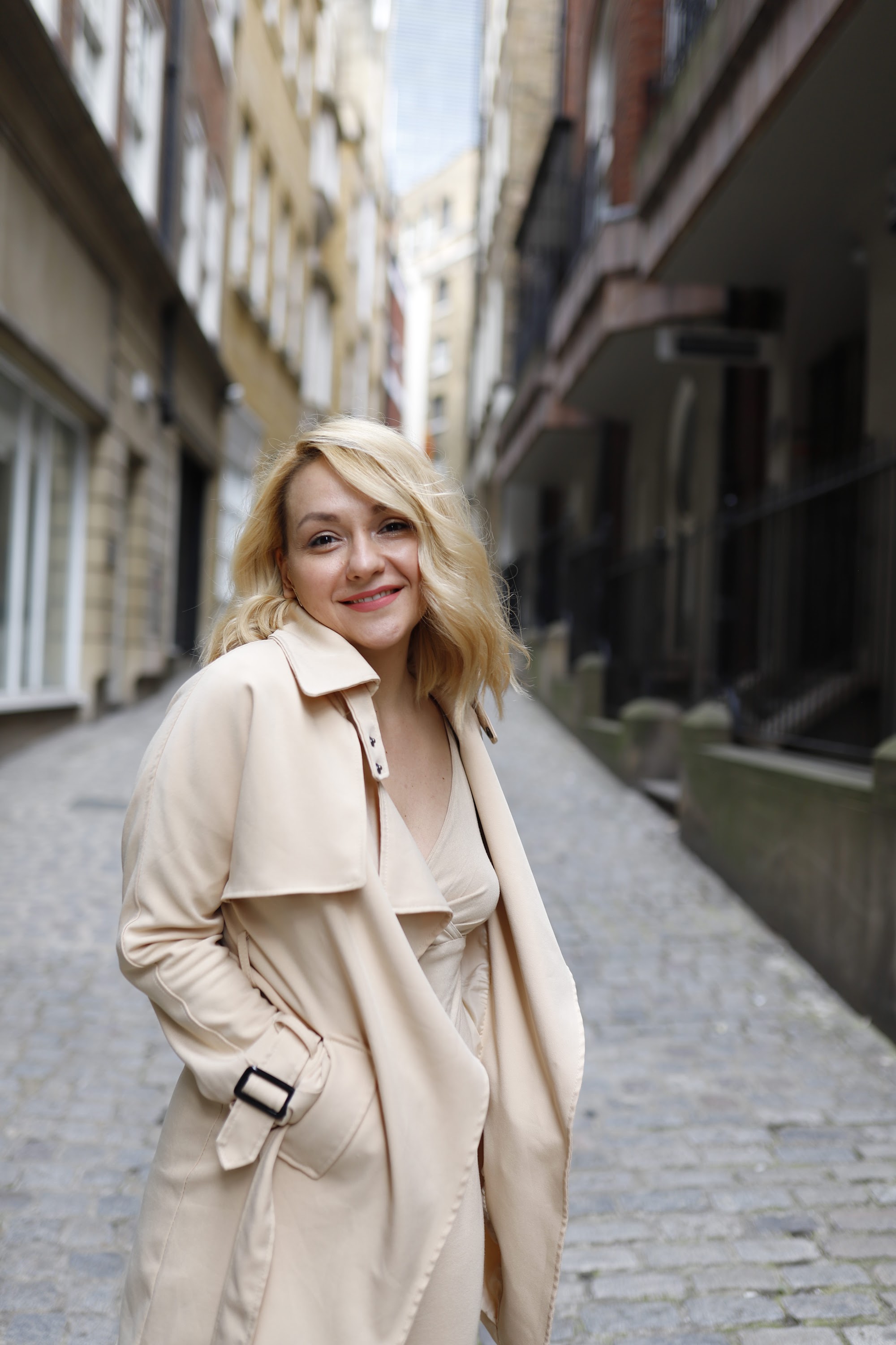 Blonde Woman in Beige Trench Coat on Cobblestone Alley