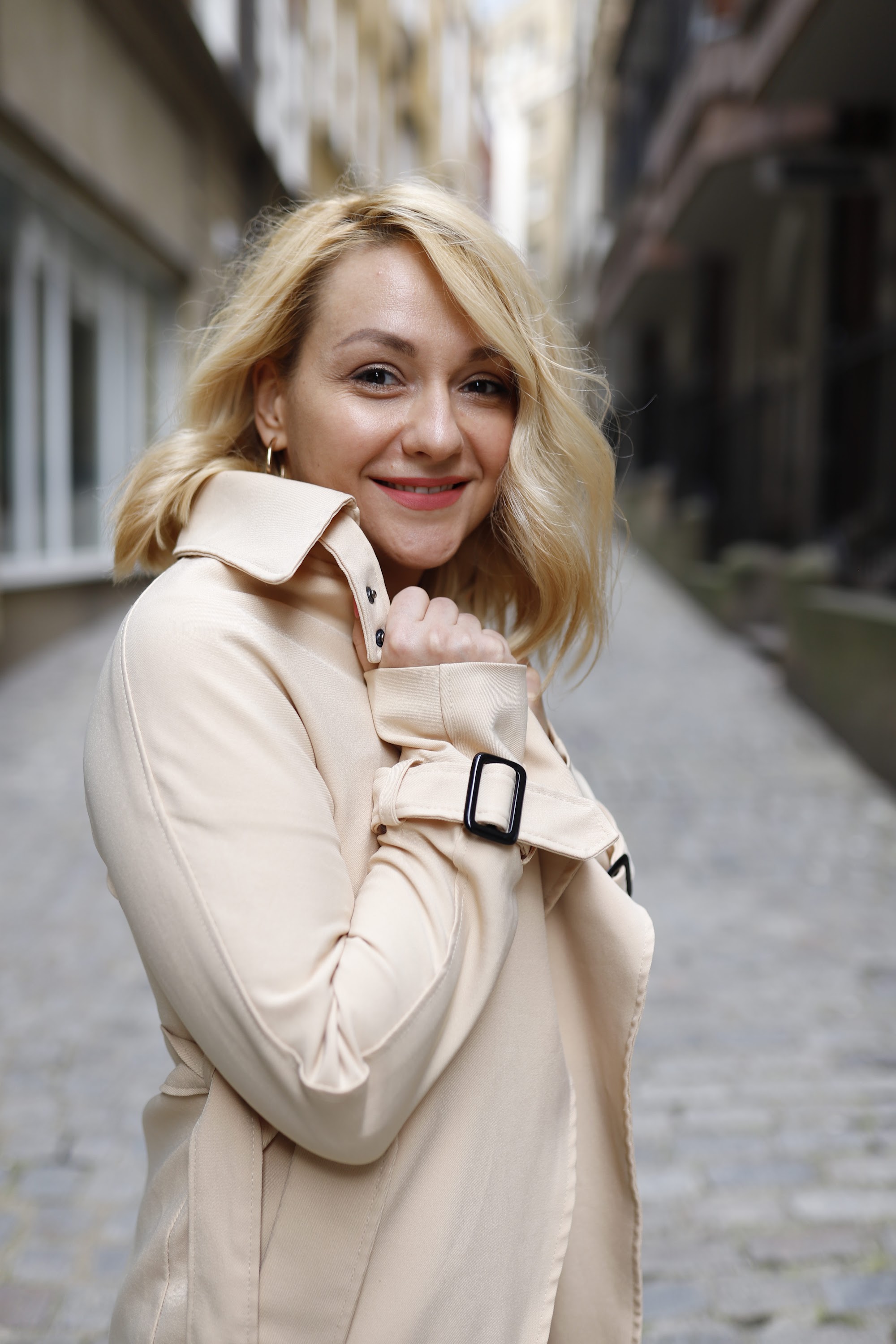 Blonde Woman in Beige Trench Coat on Cobblestone Alley