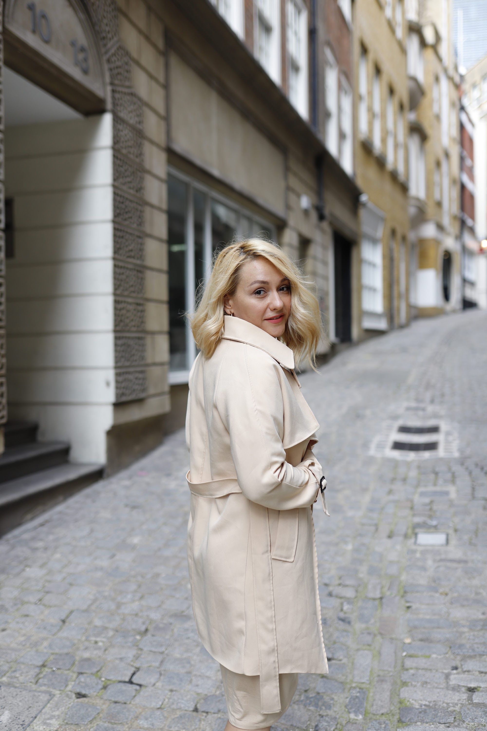 Blonde Woman in Beige Trench Coat on Cobblestone Alley
