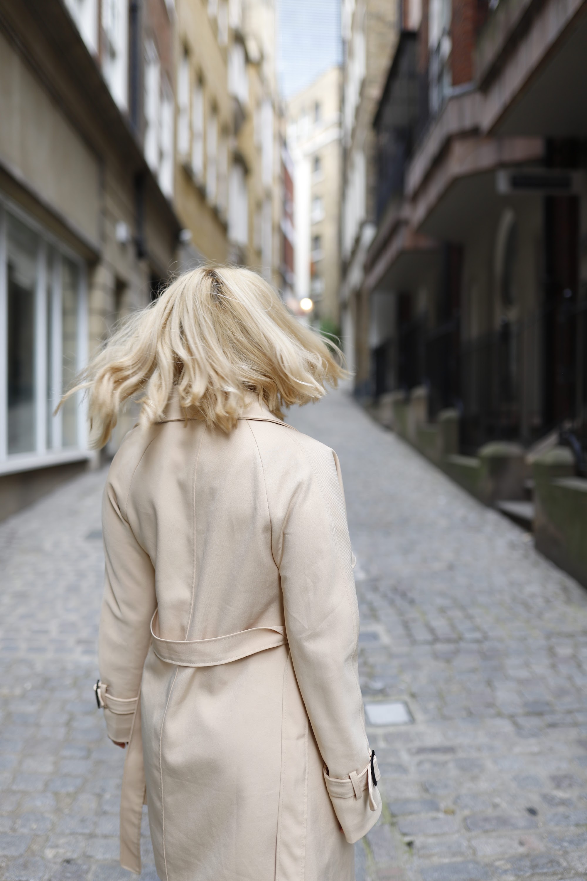 Blonde Woman in Beige Trench Coat on Cobblestone Alley