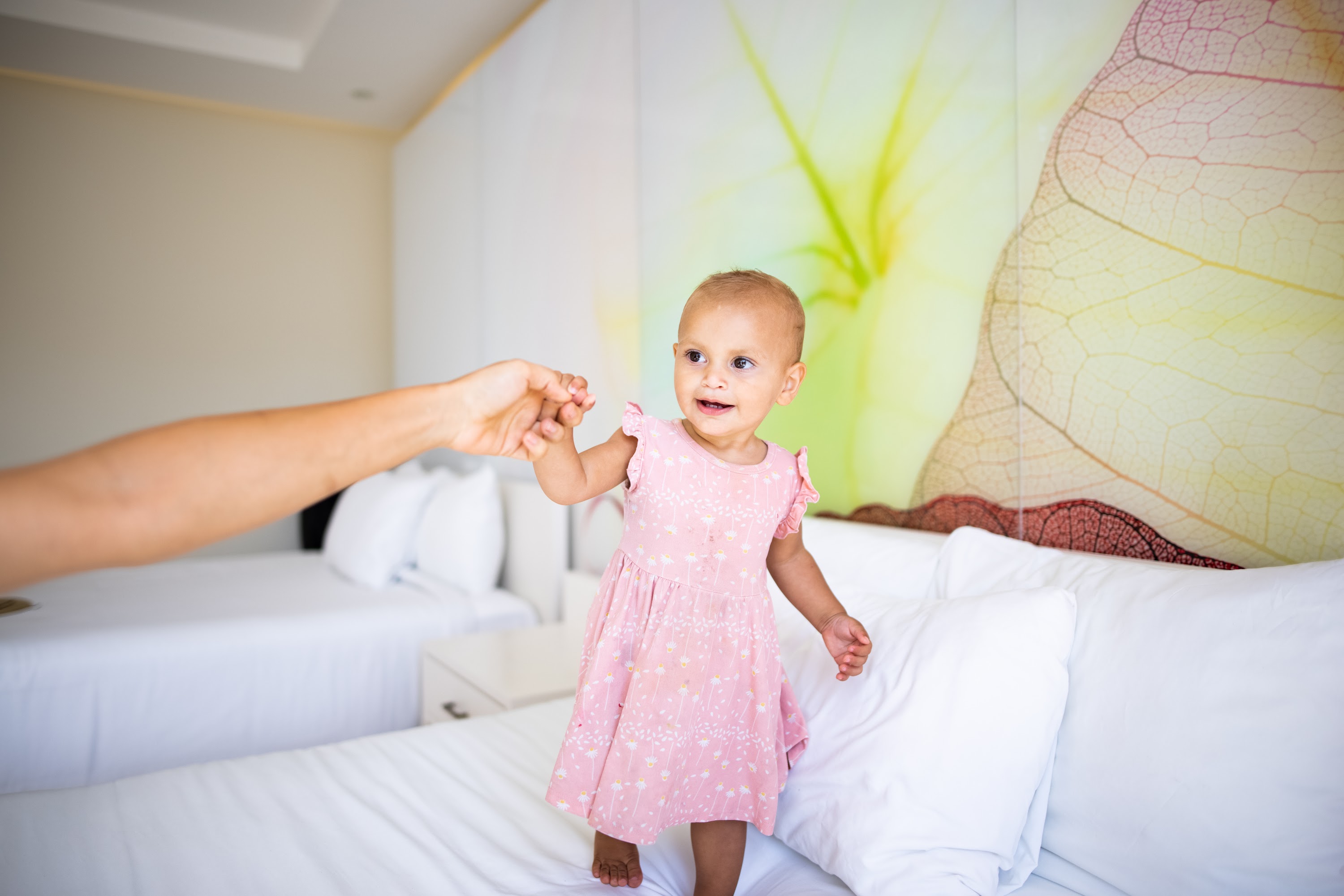 Baby in Pink Dress Holding Adult's Hand on Bed
