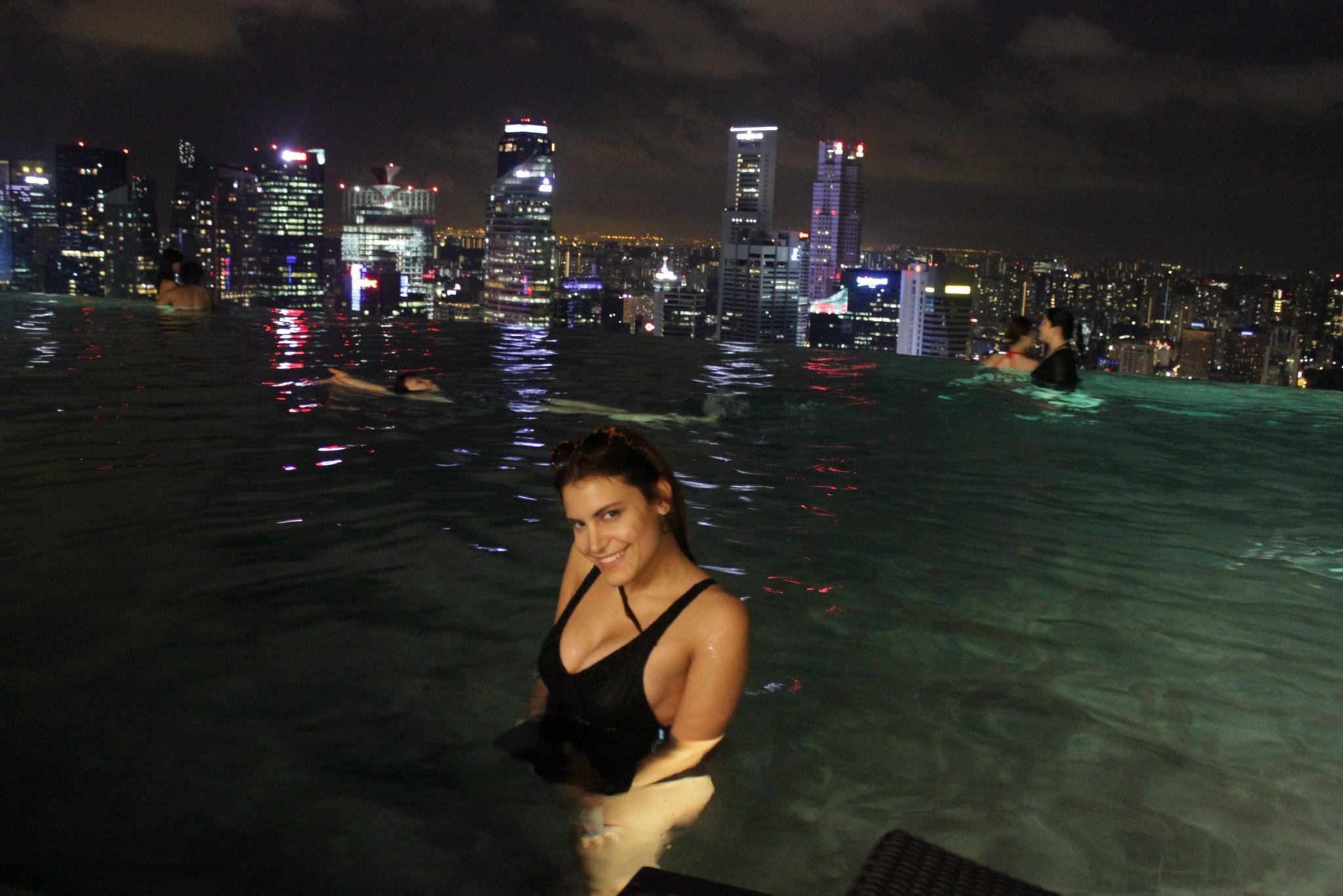 Woman smiling in rooftop infinity pool at night with city skyline
