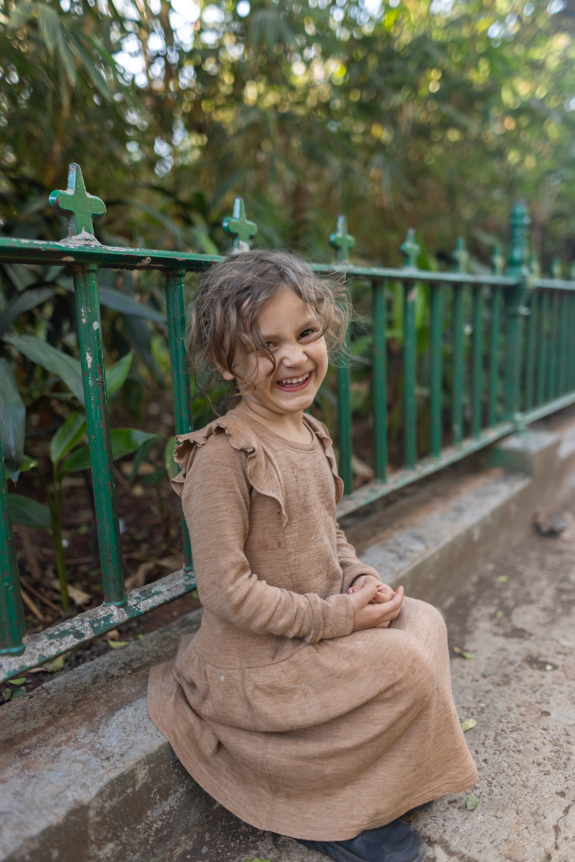 Smiling Girl in Brown Dress Sitting by Green Fence